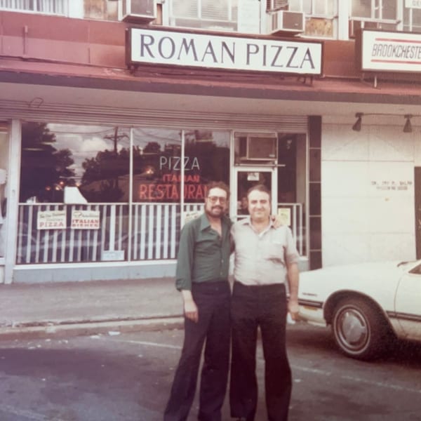 Two men stand outside Roman Pizza & Restaurant, with a white car parked nearby.