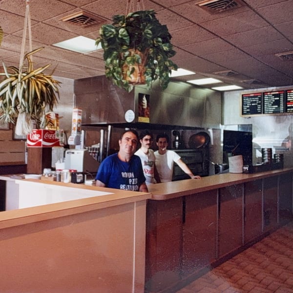 Three men, likely Roman Pizza & Restaurant staff, stand behind a counter in a retro kitchen.