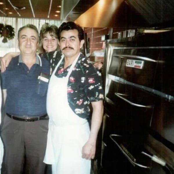 Three Roman Pizza & Restaurant staff members, two men and one woman, smiling in the kitchen.
