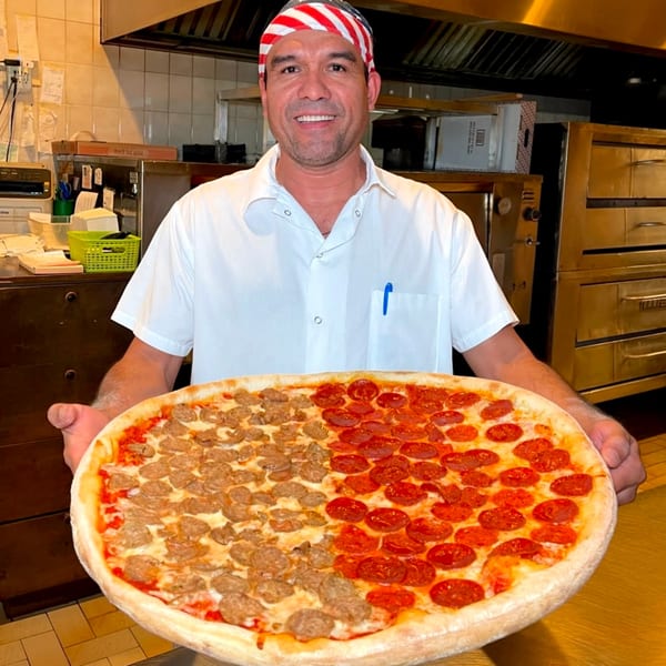 A smiling Roman Pizza chef holds a large half-pepperoni, half-sausage pizza in a kitchen.
