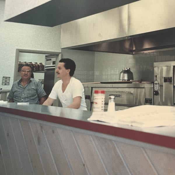 Two men behind the counter of Roman Pizza & Restaurant, with pizza ovens in the background.