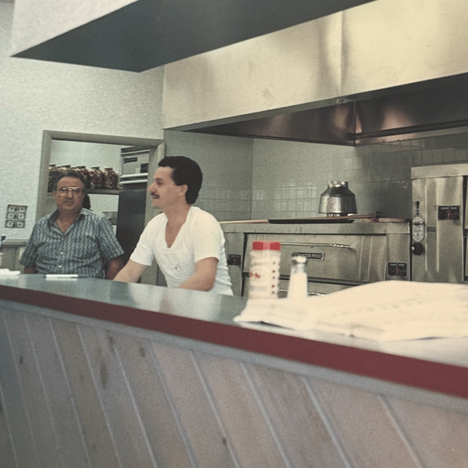 Two men behind the counter of Roman Pizza & Restaurant, with pizza ovens in the background.
