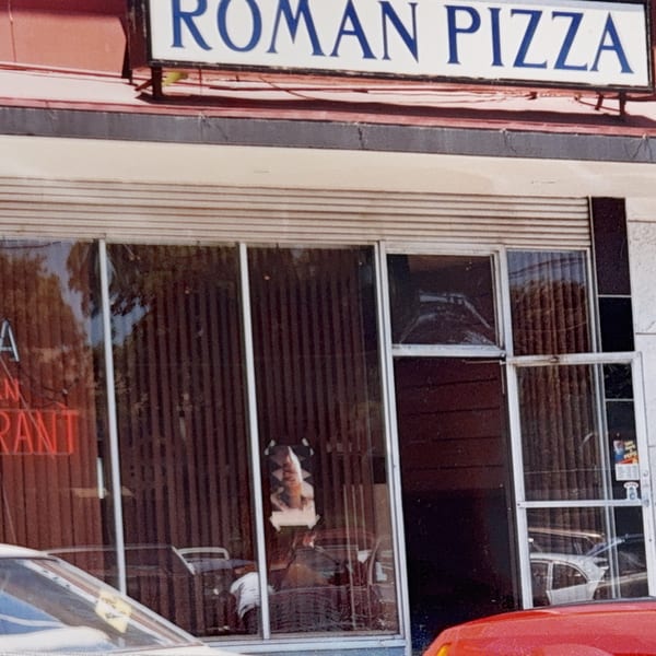 Exterior of Roman Pizza & Restaurant with a red neon sign visible through the window.