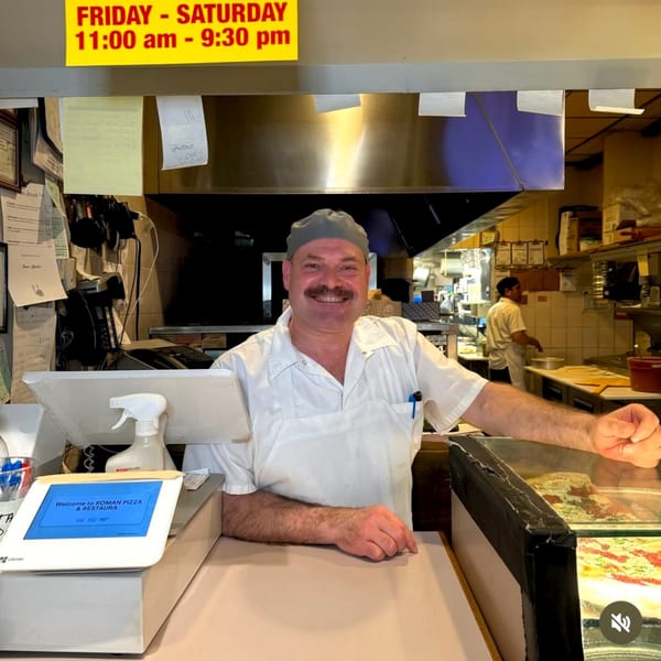 Smiling Roman Pizza & Restaurant chef in a white uniform and gray cap behind the counter.