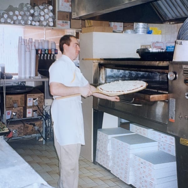 A Roman Pizza chef slides a raw pizza into a large commercial oven in a busy kitchen.