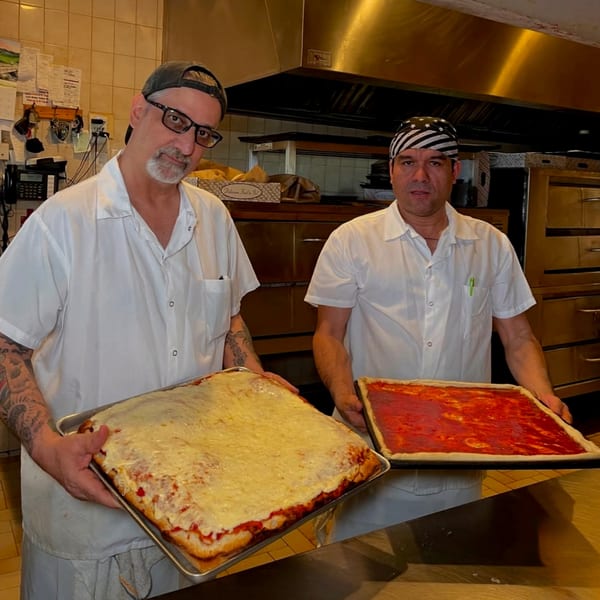 Two Roman Pizza chefs in a kitchen holding large rectangular pizzas, one with cheese, one with sauce.