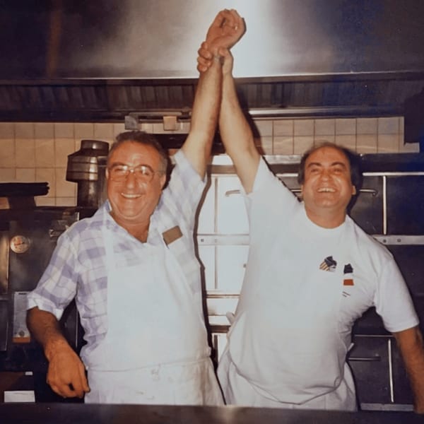 Two smiling Roman Pizza chefs in aprons, raising clasped hands in celebration in a kitchen.