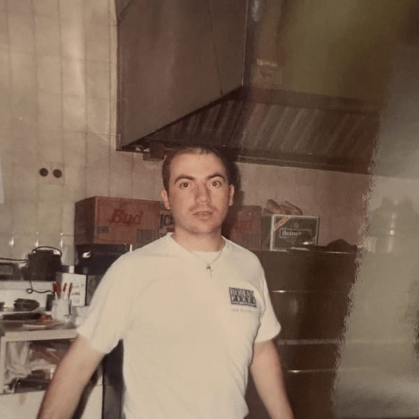 A man in a white Roman Pizza & Restaurant t-shirt stands in a kitchen with beer boxes.