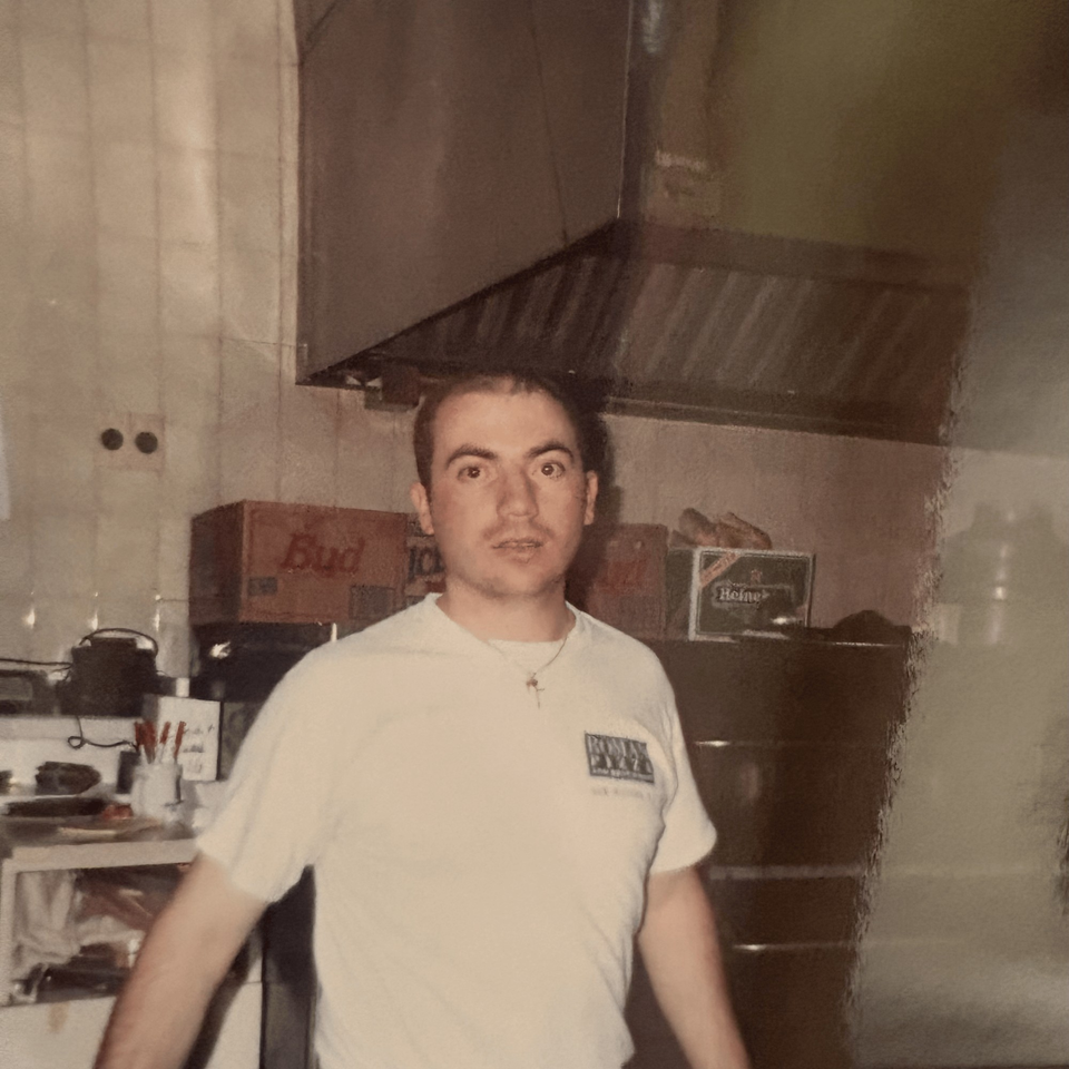 A man in a white Roman Pizza & Restaurant t-shirt stands in a kitchen with beer boxes.