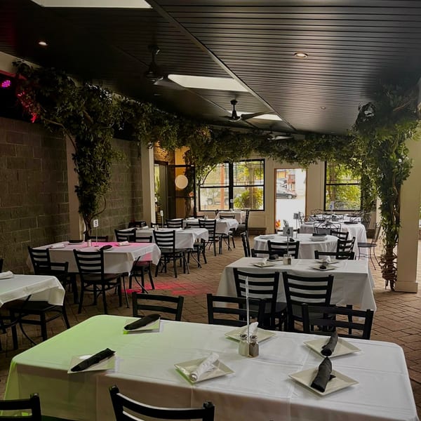 Outdoor dining area at Roman Pizza & Restaurant with white-clothed tables, black chairs, and overhead greenery.