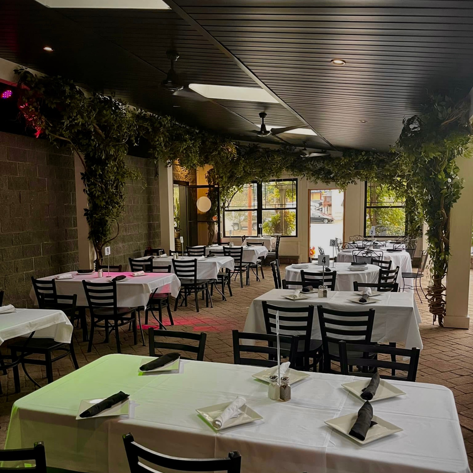 Outdoor dining area at Roman Pizza & Restaurant with white-clothed tables, black chairs, and overhead greenery.