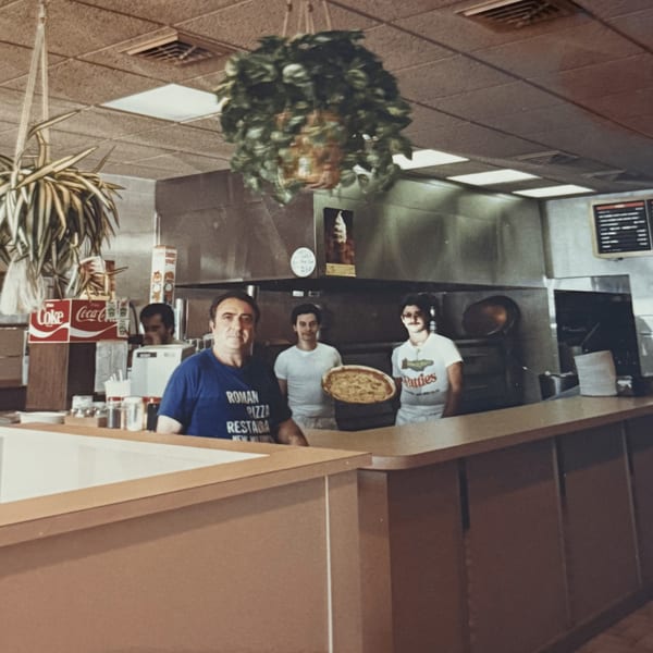 Three men, two holding pizzas, stand behind the counter of Roman Pizza & Restaurant.