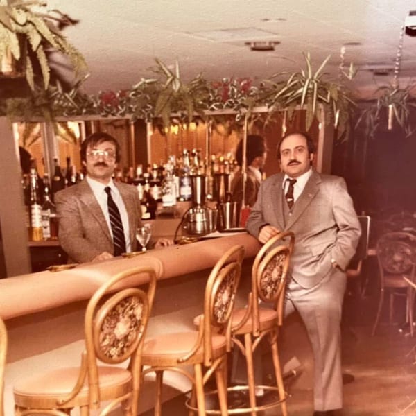 Two men in suits stand behind and beside a bar with bottles and hanging plants.