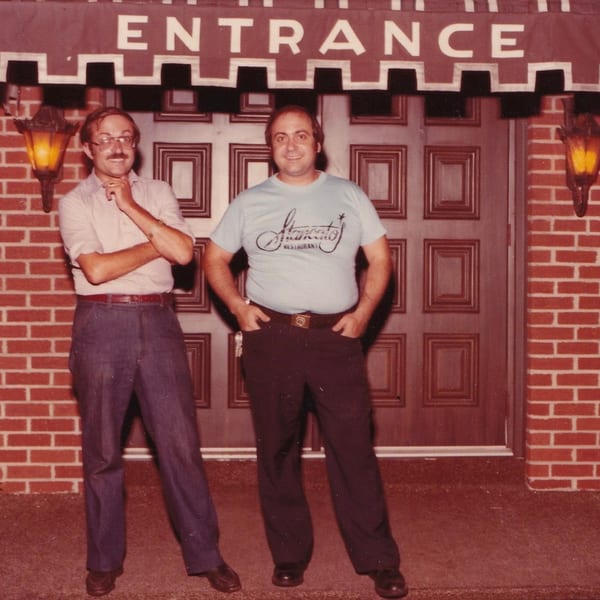 Two men standing outside Roman Pizza & Restaurant, one wearing a branded shirt.