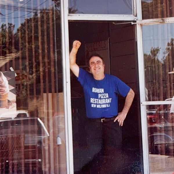 Smiling man in a blue Roman Pizza Restaurant t-shirt stands in a doorway.