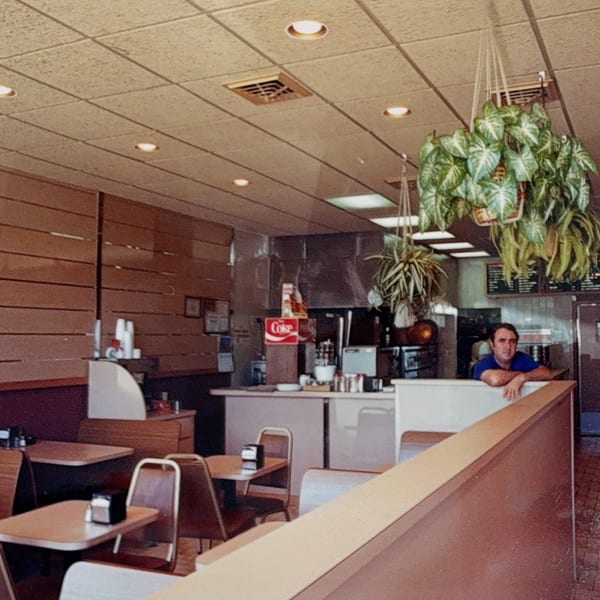 A man leans on a counter in a Roman Pizza & Restaurant dining area with tables and hanging plants.