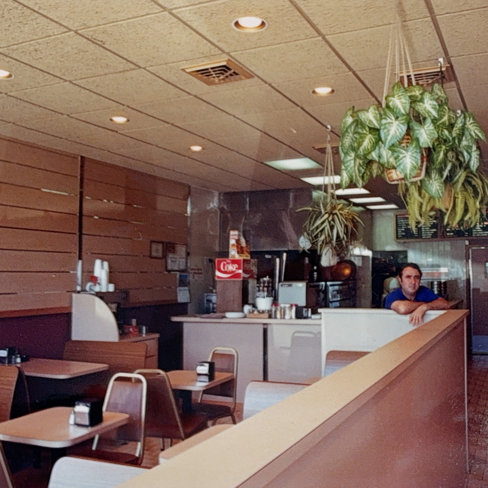 A man leans on a counter in a Roman Pizza & Restaurant dining area with tables and hanging plants.