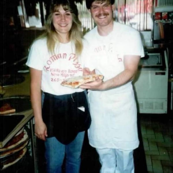 Two smiling Roman Pizza employees, a woman and a man, holding a slice of pizza.