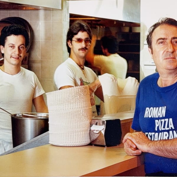 Three men, two smiling, stand behind a counter in the Roman Pizza & Restaurant kitchen.