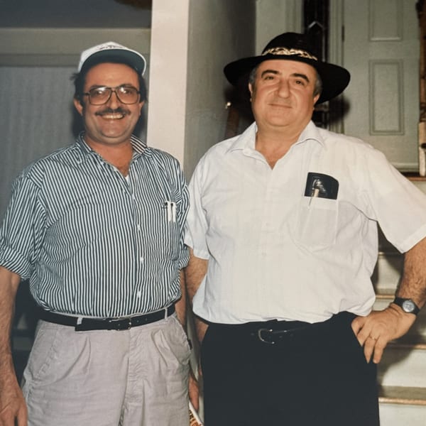 Two men, one in a striped shirt and cap, the other in a white shirt and hat, smiling.