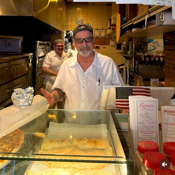 A Roman Pizza & Restaurant chef smiles behind a counter displaying pizzas and calzones.