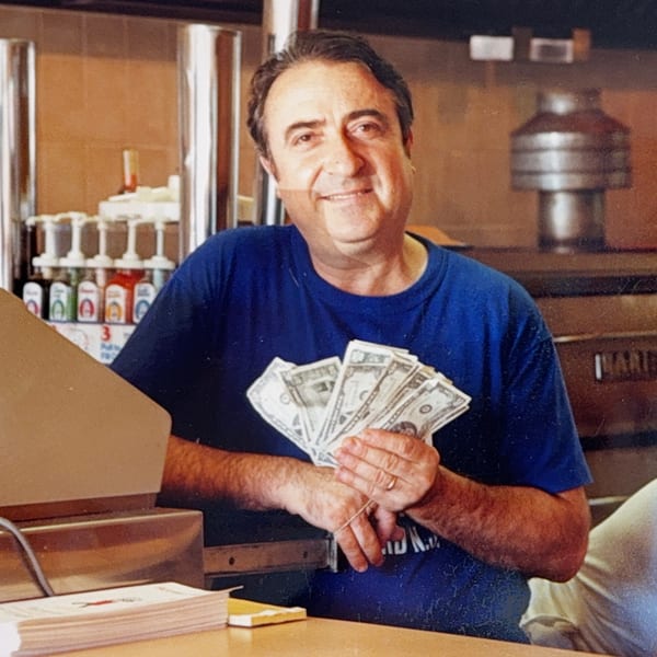 Smiling Roman Pizza owner holding a fan of cash behind the counter.