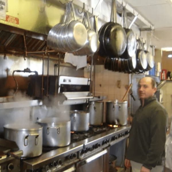 Chef standing in a busy Roman Pizza kitchen with steaming pots on the stove and hanging pans.
