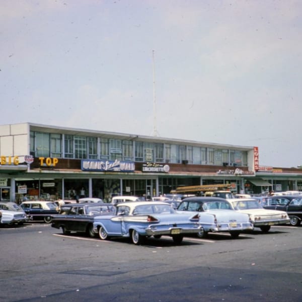 A vintage street view of a shopping center with classic cars parked in front of various businesses.