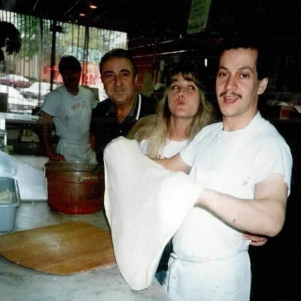 A Roman Pizza chef stretches pizza dough while three people watch in the background.