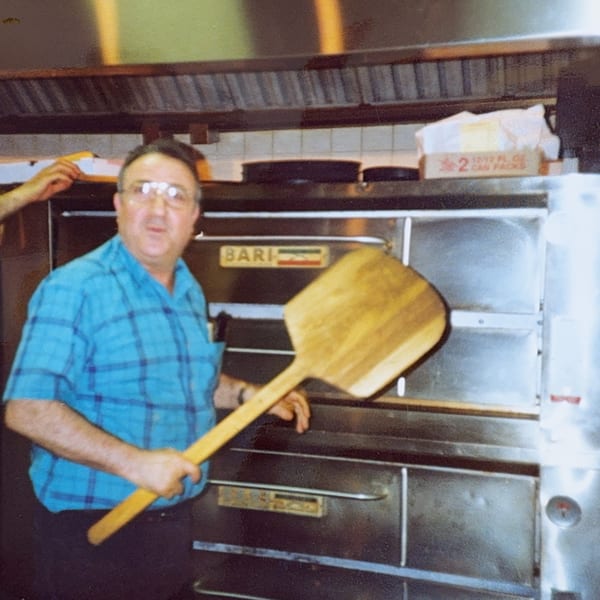 A man in a blue plaid shirt holds a pizza peel in front of a Bari pizza oven.