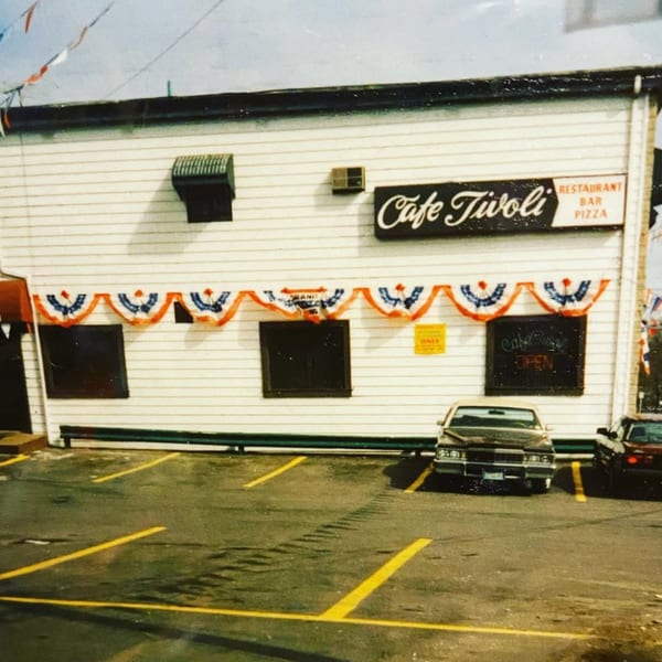 Exterior of Cafe Tivoli, a white building with patriotic bunting, a sign, and cars in the parking lot.