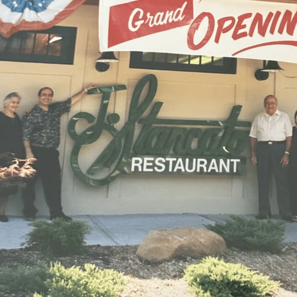 Three people standing outside Roman Pizza & Restaurant under a "Grand Opening" banner.