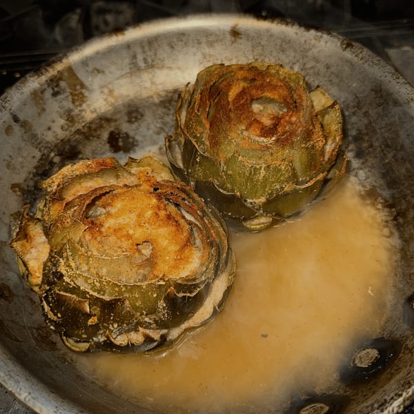 Two golden-brown fried artichokes in a pan with a light sauce, a Roman Pizza & Restaurant appetizer.