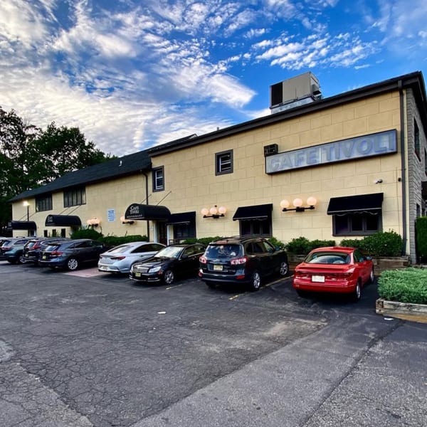Exterior of Roman Pizza & Restaurant, a yellow building with black awnings and a parking lot full of cars.
