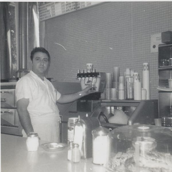 A black and white photo of a man in a white shirt and apron standing behind a counter with a soda fountain.