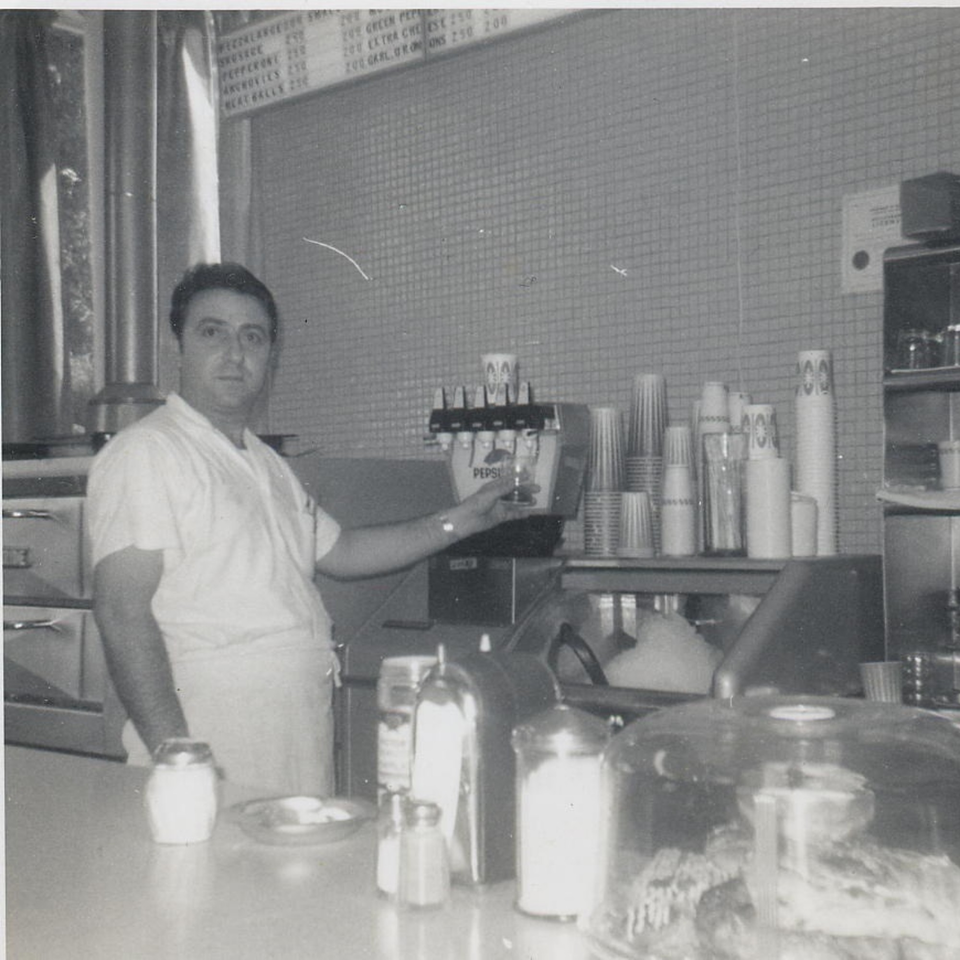 A black and white photo of a man in a white shirt and apron standing behind a counter with a soda fountain.