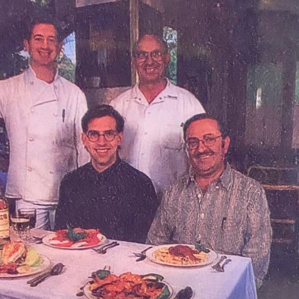 Four smiling men, two chefs and two diners, seated at a table with Italian dishes.