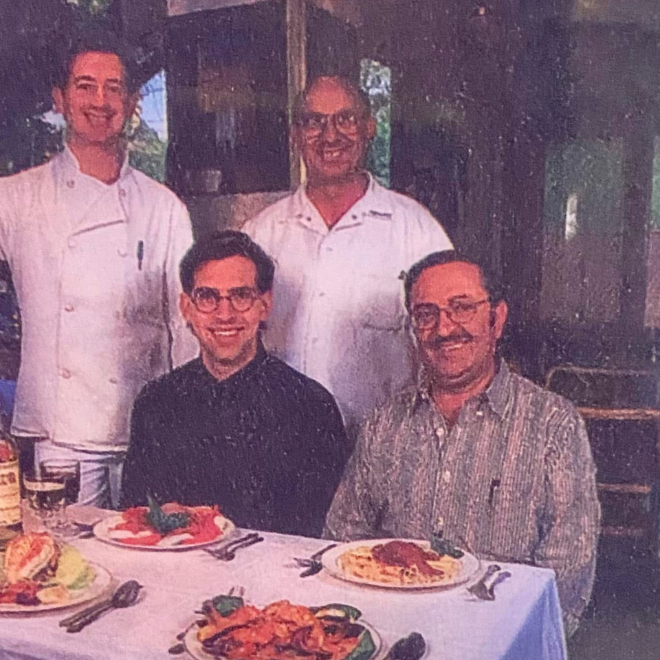 Four smiling men, two chefs and two diners, seated at a table with Italian dishes.