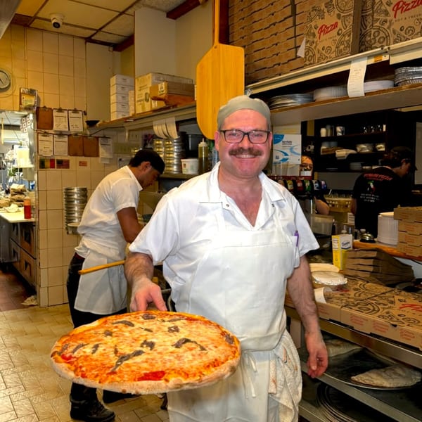 Smiling Roman Pizza chef holding a freshly baked anchovy pizza in a busy kitchen.