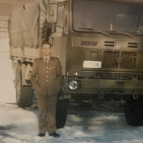 A man in a military uniform stands in front of a large military truck on a snowy ground.