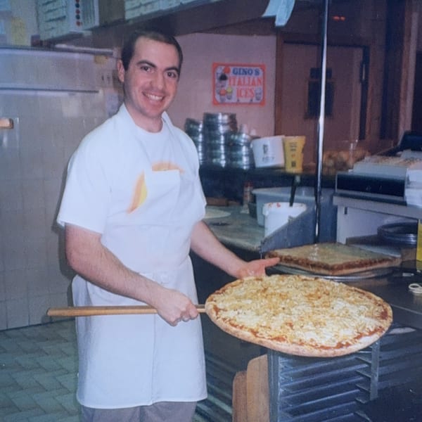 Smiling Roman Pizza chef in a white apron holding a large round pizza on a peel.
