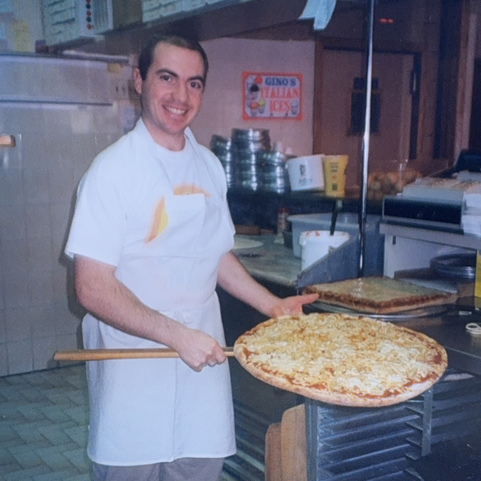 Smiling Roman Pizza chef in a white apron holding a large round pizza on a peel.