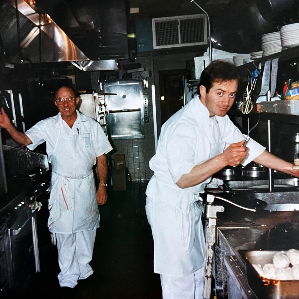 Two chefs in white uniforms working in a busy Roman Pizza & Restaurant kitchen.